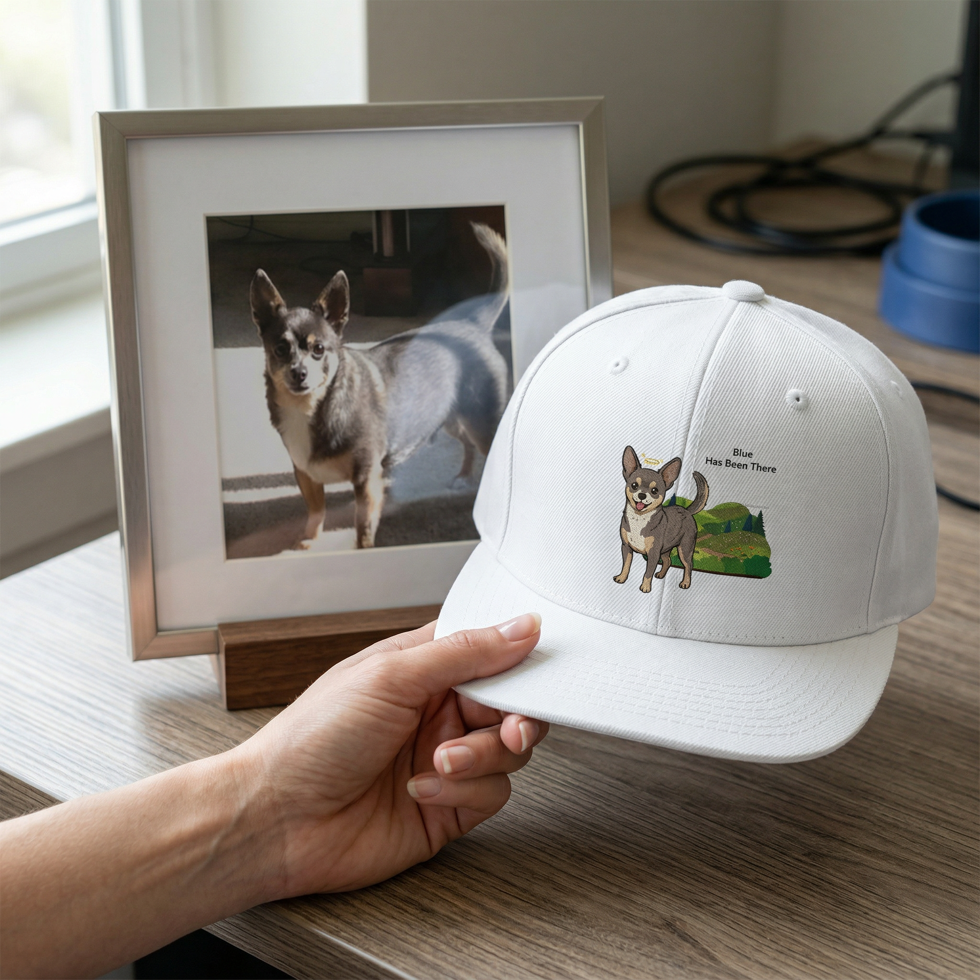 A snapback hat with a custom design of a Chihuahua dog who crossed the rainbow bridge and a wildflower trail background. The hat is held next to the photo of the dog as a memorial gift.