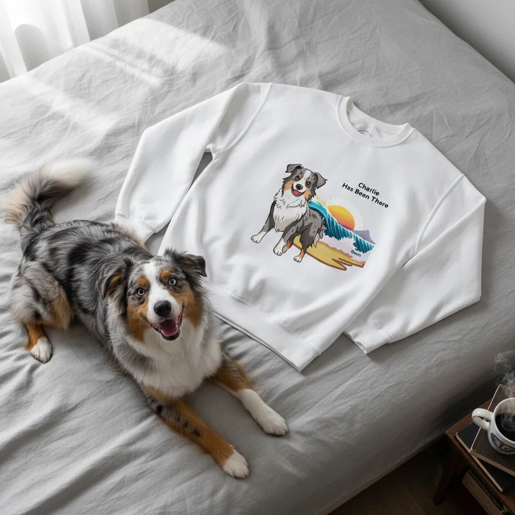 Personalized sweatshirt with a dog illustration, dog name, "Has Been There" badge, and beach illustration, lying on a bed with an Australian Shepherd next to it. Outdoor Adventure Series.