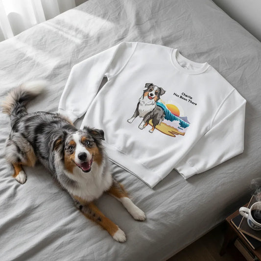 Personalized sweatshirt with a dog illustration, dog name, "Has Been There" badge, and beach illustration, lying on a bed with an Australian Shepherd next to it. Outdoor Adventure Series.