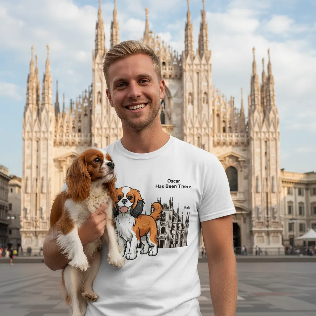 Male wearing a personalized t-shirt with a dog illustration, dog name, "Has Been There" badge, and Italy illustration, holding a King Charles Spaniel dog in front of the Duomo di Milano. World Travel Series.
